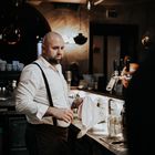 Ein Mann mit Glatze und Bart arbeitet in einer Bar, trägt ein weißes Hemd mit schwarzen Hosenträgern und trocknet Gläser ab.