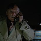 Keine erkennbaren Texte. Mann in Trenchcoat telefoniert in einem dunklen Raum, besorgt dreinblickend.