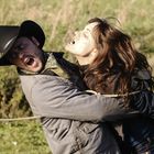 Ein Mann mit Cowboyhut hält eine Frau fest. Beide haben erschrockene Gesichtsausdrücke, und sie stehen auf einem Feld.