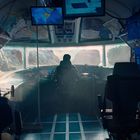 Verschiedene Monitore mit Diagrammen und Karten in einem futuristischen Kontrollraum; eine Person sitzt in der Mitte.