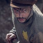 Es gibt keine sichtbaren Texte. Ein Mann mit Mütze und dreckigem Gesicht schaut direkt, trägt einen Pulli mit gelbem Fleck.