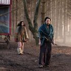 Zwei Personen in traditioneller Kleidung stehen in einem Wald mit einem hölzernen Wagen im Hintergrund.