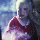 Ein blondes Mädchen in roter Jacke steht vor nebligem Hintergrund, ihr Haar wird hochgeweht.