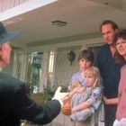 Ein alter Mann in Hut spricht eine Familie auf einer Veranda an: zwei Erwachsene, zwei Kinder, die ein Kuscheltier halten.