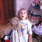 Ein Kind mit langem blondem Haar hält eine Puppe. Es trägt ein T-Shirt mit bunten Früchten. Im Hintergrund ein Tisch und eine Puppe.