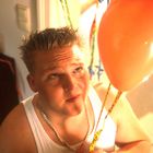 Ich kenne die Texte nicht. Ein Mann blickt nach oben zu einem orangenen Ballon. Er trägt ein weißes Unterhemd und eine Kette.