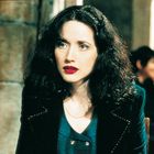 Es gibt keinen Text. Eine Frau mit dunklem, lockigem Haar und rotem Lippenstift trägt eine dunkle Jacke mit goldenen Details.