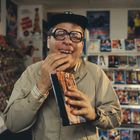 Ein Mann mit großer Brille lächelt, hält eine VHS-Kassette. Im Hintergrund Regale voller Videos.