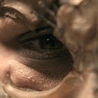 Ein Auge blickt durch eine ungleichmäßige, maskenartige Struktur aus Wachs oder Ton. Es wirkt geheimnisvoll und intensiv.