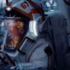 Zahl "63" auf Helm. Person in Raumanzug mit leuchtendem Visier, digitalen Anzeigen sichtbar. Hintergrund unscharf.