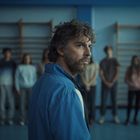 Ein Mann im blauen Trainingsanzug steht in einer Turnhalle; unscharfe Gruppe im Hintergrund.