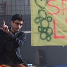 Text: "SPIEG L'AM". Mann mit Brille und kariertem Sakko sitzt, zeigt Handgeste, im Hintergrund ein gelbes Banner.