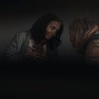Eine Frau mit besorgtem Blick steht einer Person mit gebeugter Haltung gegenüber. Die Szene ist dunkel und intim.