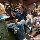 „CINX“ auf einem T-Shirt. In einem Wohnzimmer sitzen Menschen, eine Frau tanzt im Schoß eines Mannes, es herrscht Partystimmung.