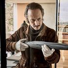 Ein Mann mit brauner Jacke und Handschuhen hält konzentriert einen geschlossenen Regenschirm in einem modernen Raum.