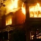 Ein brennendes zweigeschossiges Backsteinhaus, Flammen lodern aus Fenstern, Rauch steigt auf, ein dramatisches Feuer.