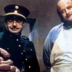 Zwei Männer betrachten skeptisch einen Turm aus großen Würsten, einer in Uniform mit Mütze, der andere trägt eine Schürze.