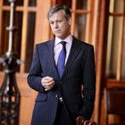 Kein sichtbarer Text vorhanden. Ein Mann im Anzug steht in einem Raum mit Holztüren und großen Fenstern.