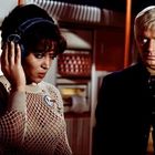 Eine Frau mit Kopfhörern im Netzpullover steht neben einem Mann. Beide wirken konzentriert in einem futuristischen Raum.