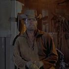 Ein Mann mit Cowboyhut steht in einem Stall neben Scheunentoren, umgeben von Holzwänden und einem Hufeisen an der Wand.