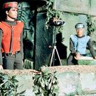 Zwei Puppen in farbigen Uniformen, eine mit Fernglas auf Stativ, von Pflanzen umgeben, wirken wachsam in einer Modelllandschaft.