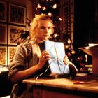 Eine Frau bindet ein Paket mit blauem Papier, in einem festlich beleuchteten Raum mit Weihnachtsbaum im Hintergrund.