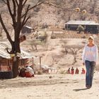 Eine Frau in weißem Oberteil und Jeans läuft durch eine trockene Landschaft mit Bäumen und Hütten im Hintergrund.