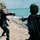 Zwei Männer fechten auf einer felsigen Küste, einer hat einen Helm, der andere trägt einen schwarzen Mantel. Meer im Hintergrund.