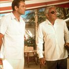 Zwei Männer in Sommerkleidung stehen nebeneinander auf einer Terrasse. Einer trägt eine Augenklappe, im Hintergrund Palmen.