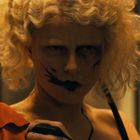 Eine Person mit blonden Locken und dunklem Gesichtsschmuck blickt ernst, mit dramatischem Make-up und einem orangefarbenen Kleid.