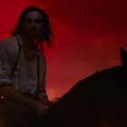 Ein Mann auf einem Pferd vor einem feurig roten Hintergrund, trägt ein weißes Hemd und hat lange Haare.