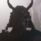 Ein Krieger mit dunkler Rüstung und roten Augen, trägt einen verzierten Helm mit Hörnern, umgeben von Dämmerlicht.