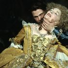 Ich kann den Text nicht lesen. Eine Frau in altmodischem gelbem Kleid wird von einem Mann im blauen Mantel am Hals gehalten.