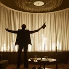 Eine Person steht mit ausgestreckten Armen vor leuchtendem Vorhang, hält einen Hut. Lounge-Atmosphäre mit Sesseln und Tisch.