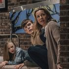 Ein kleines Mädchen, ein Junge und eine Frau kauern sich ängstlich hinter einer Backsteinmauer.