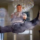 Ein Mann in Aktion schwebt, Beine gespreizt, mit entschlossenem Gesichtsausdruck und Defibrillator, umgeben von Rauch.