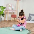 Ein Kind umarmt ein Plüschtier und sitzt auf einem grünen Teppich in einem hellen Zimmer mit Pflanzen.