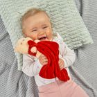 Ein lachendes Baby hält ein rotes Kuscheltuch in Form eines Bären. Es liegt auf einem weichen, hellen Kissen.