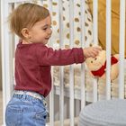 Ein Kleinkind in rotem Pullover und Jeans spielt mit einem kleinen, flauschigen Teddybären an einem weißen Gitterbett.