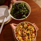 Ein Teller mit gemischtem Grün wird von einer Hand gehalten; darunter eine Pfanne mit Reis, Garnelen und Gemüse auf einem Holztisch.