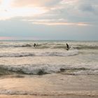 Zwei Surfer reiten in Neoprenanzügen auf kleinen Wellen unter bewölktem Himmel, während die Sonne am Horizont untergeht.