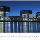 Text: "Kranhäuser" und "3". Drei moderne, rechteckige Gebäude am Fluss bei Nacht, beleuchtet, spiegeln sich im Wasser.