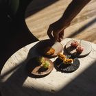 Kein Text vorhanden. Eine Hand greift nach einer von vier kunstvoll angerichteten Speisen auf kleinen Tellern, beleuchtet durch Sonnenlicht auf einem Tisch.
