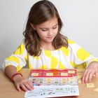 Zahlenkarten von 1 bis 7. Ein Mädchen in gelb-weißem Kleid spielt mit einem Zahlen- und Symbolspiel auf einem Tisch.