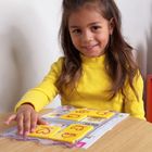 Auf gelben Karten befinden sich rote Buchstaben. Ein lächelndes Mädchen in gelbem Pullover spielt damit an einem Tisch.