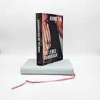 Aufrecht stehender Roman "Die Liebeshungrigen" von Karine Tuil. Darauf Hände mit Ringen, darunter ein helles Buch.