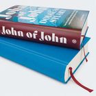 "John of John" steht auf einem Buchrücken in Weiß auf Rot. Zwei Bücher sind gestapelt, eines blau, das obere mit Schutzumschlag.