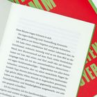 Text auf einer Buchseite beginnt mit: "Diese Räume tragen Schmerz in sich. Sie tragen Hoffnung, Leid, Verzweiflung, Zuversicht." Im Hintergrund rote Buchumschläge mit grünen Buchstaben.