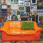 Grünes Kissen mit roter Schrift "Familie Montes" auf einer orangefarbenen Couch vor einer Wand mit vielen Kunstwerken.