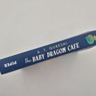 "A. T. QURESHI, The BABY DRAGON CAFE" steht auf einem blauen Buchrücken mit einem kleinen Drachen-Illustration.